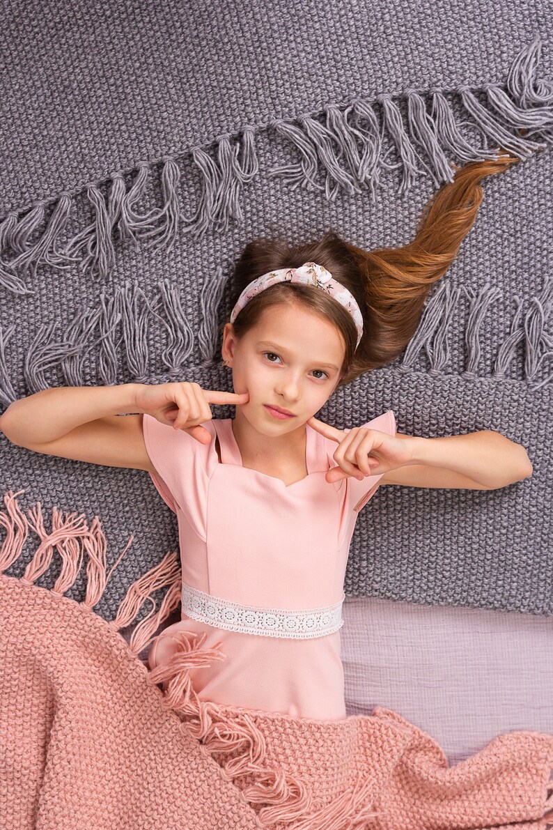 May include: A young girl with long brown hair wearing a pink dress with a white lace trim lies on a gray and pink blanket. She is looking at the camera and has her fingers on her cheeks.