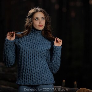 May include: A woman wearing a blue knitted sweater with a textured pattern. The sweater has a high neckline and long sleeves. The woman is standing in a dark forest setting.