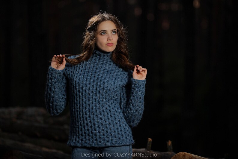 May include: A woman wearing a blue knitted sweater with a textured pattern. The sweater has a high neckline and long sleeves. The woman is standing in a dark forest setting.