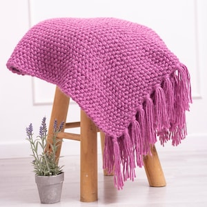 May include: A soft, pink knitted throw blanket with fringe on the edges. The blanket is draped over a wooden stool.