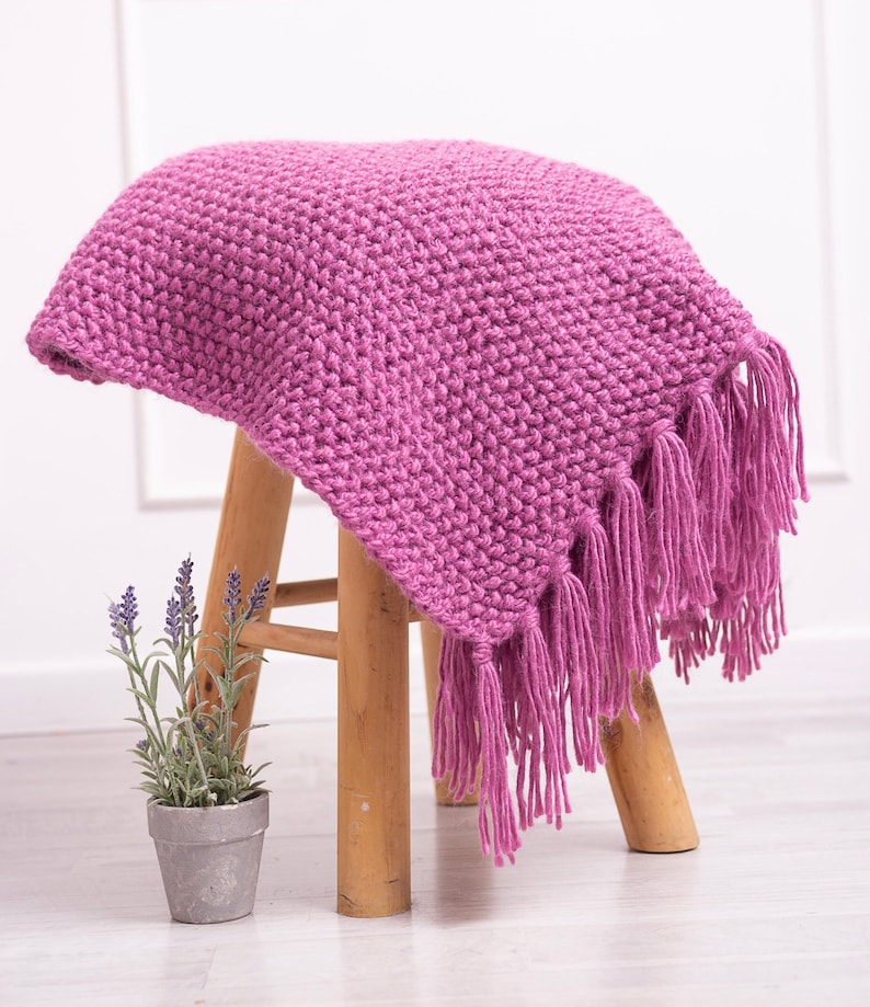 May include: A soft, pink knitted throw blanket with fringe on the edges. The blanket is draped over a wooden stool.