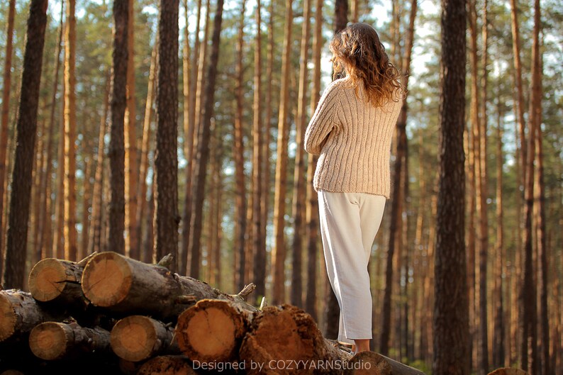 May include: A woman wearing a cream-colored sweater and white pants stands on a pile of logs in a forest. The trees are tall and slender, and the sun is shining through the branches.