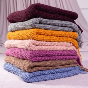 May include: A stack of seven knitted blankets in various colors, including purple, pink, brown, blue, yellow, gray, and burgundy. The blankets are folded and stacked on top of each other, creating a cozy and inviting display.