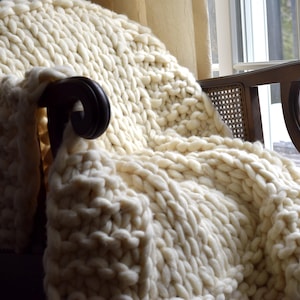 May include: A white chunky knit throw blanket draped over a wooden armchair with a woven cane back.