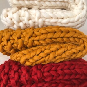 May include: Three chunky knit blankets stacked on top of each other. The top blanket is white, the middle blanket is mustard yellow, and the bottom blanket is red.