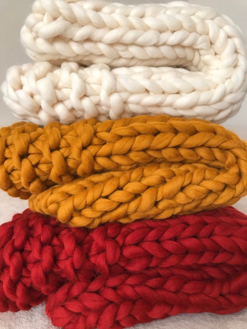May include: Three chunky knit blankets stacked on top of each other. The top blanket is white, the middle blanket is mustard yellow, and the bottom blanket is red.