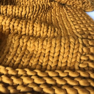 May include: A close-up of a chunky knit throw blanket in a mustard yellow color. The blanket is made of thick yarn and has a soft, fuzzy texture.