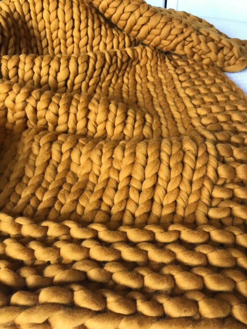 May include: A close-up of a chunky knit throw blanket in a mustard yellow color. The blanket is made of thick yarn and has a soft, fuzzy texture.