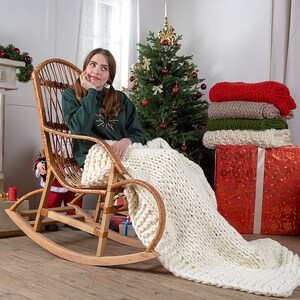 May include: A woman sits in a wicker rocking chair with a white knitted blanket draped over her legs. The chair is in front of a decorated Christmas tree and a stack of colorful blankets.