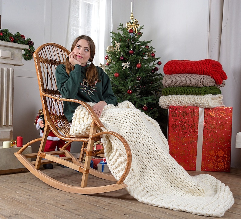 May include: A woman sits in a wicker rocking chair with a white knitted blanket draped over her legs. The chair is in front of a decorated Christmas tree and a stack of colorful blankets.
