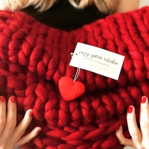 May include: A large, red, chunky knit blanket in the shape of a heart. The blanket has a small red felt heart attached to it. A tag is attached to the blanket that reads "cozy yarn studio handmade knitting with love".