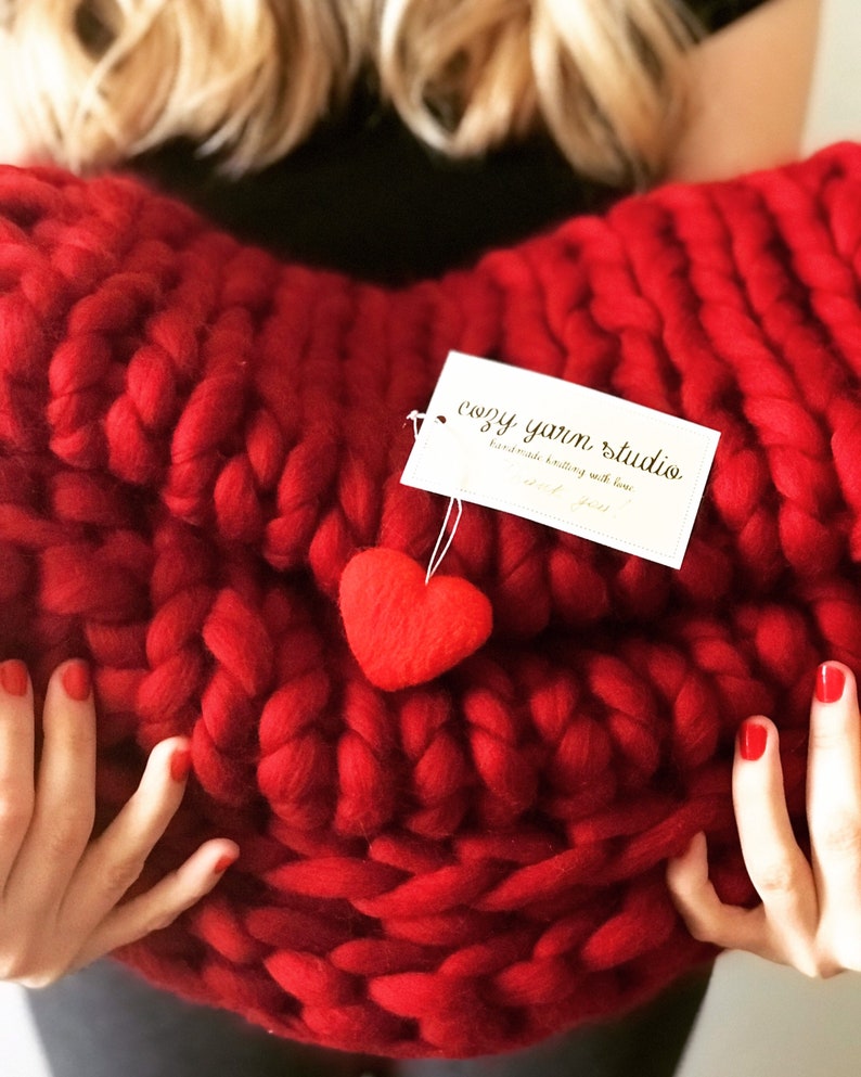 May include: A large, red, chunky knit blanket in the shape of a heart. The blanket has a small red felt heart attached to it. A tag is attached to the blanket that reads "cozy yarn studio handmade knitting with love".