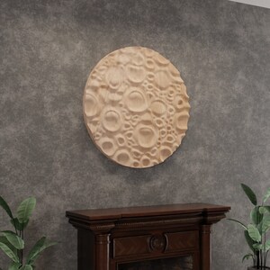 May include: A circular wooden wall sculpture with a textured surface resembling the moon. The sculpture is hanging on a grey wall above a dark wood fireplace.