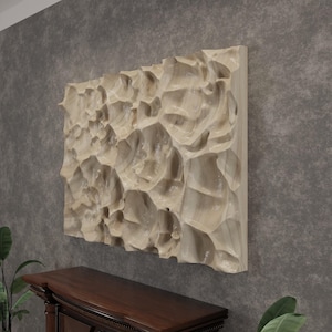 Puede incluir: Una obra de arte mural de madera clara y texturizada con un diseño orgánico tallado. La obra de arte es rectangular y está montada en una pared gris. La superficie tallada tiene un patrón irregular y fluido, creando un efecto tridimensional único.
