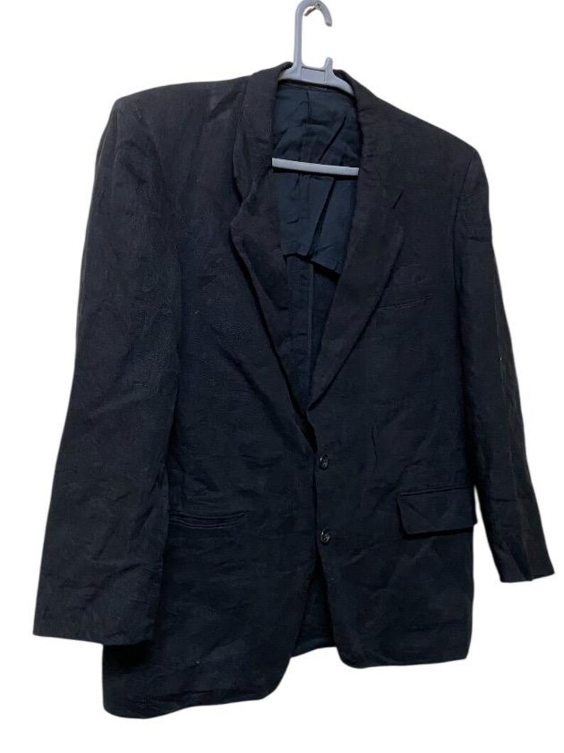 May include: A black blazer hanging on a gray hanger. The blazer has a two-button closure, a notched lapel, and a single chest pocket. The fabric appears to be a textured weave. The blazer is a classic style.