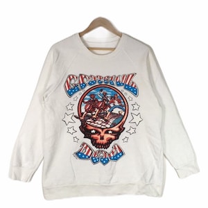 May include: White long-sleeved sweatshirt featuring a graphic print of the Grateful Dead band logo. The logo depicts a skull with a red nose and eyes, encircled by stars and a circle incorporating the band's name and an American flag design.