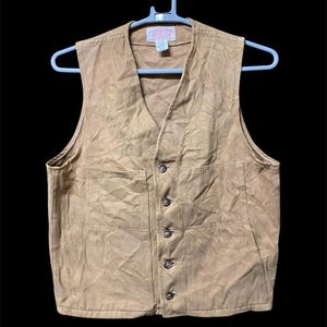 Puede incluir: Un chaleco de color beige con cuello en V y cierre de botones. El chaleco tiene dos bolsillos delanteros y est&aacute; colgado de una percha blanca. La tela parece ser un material duradero, adecuado para el trabajo o el uso informal.