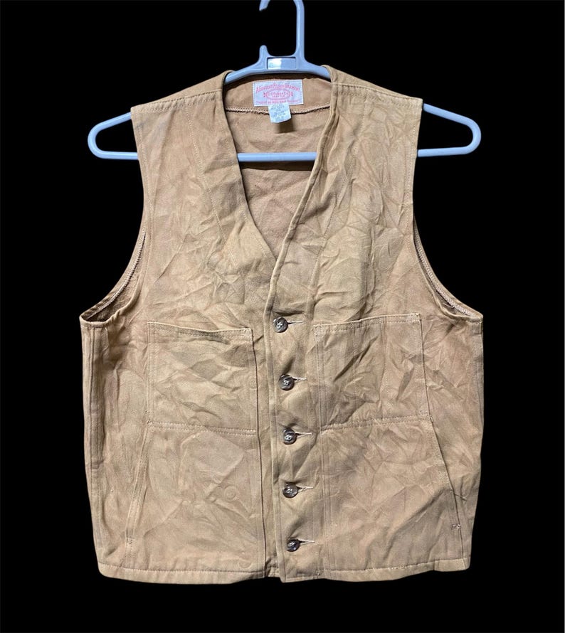 Puede incluir: Un chaleco de color beige con cuello en V y cierre de botones. El chaleco tiene dos bolsillos delanteros y est&aacute; colgado de una percha blanca. La tela parece ser un material duradero, adecuado para el trabajo o el uso informal.