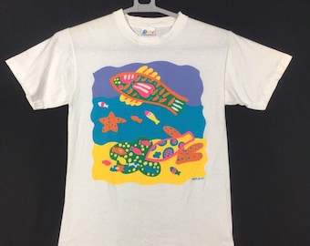 May include: White t-shirt with a colorful graphic of fish, a starfish, and a turtle in a blue and yellow ocean scene. The graphic is in a cartoon style and features bright colors.