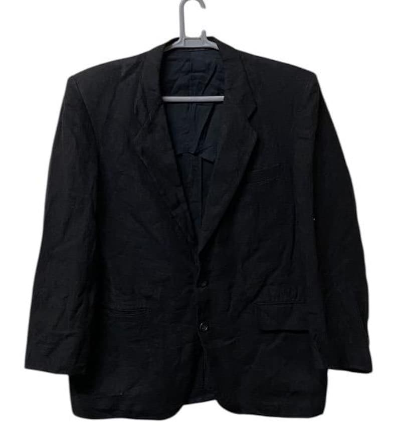 May include: A black blazer hanging on a gray hanger. The blazer has a notched lapel, two front pockets, and a two-button closure. The fabric appears to be a textured material.