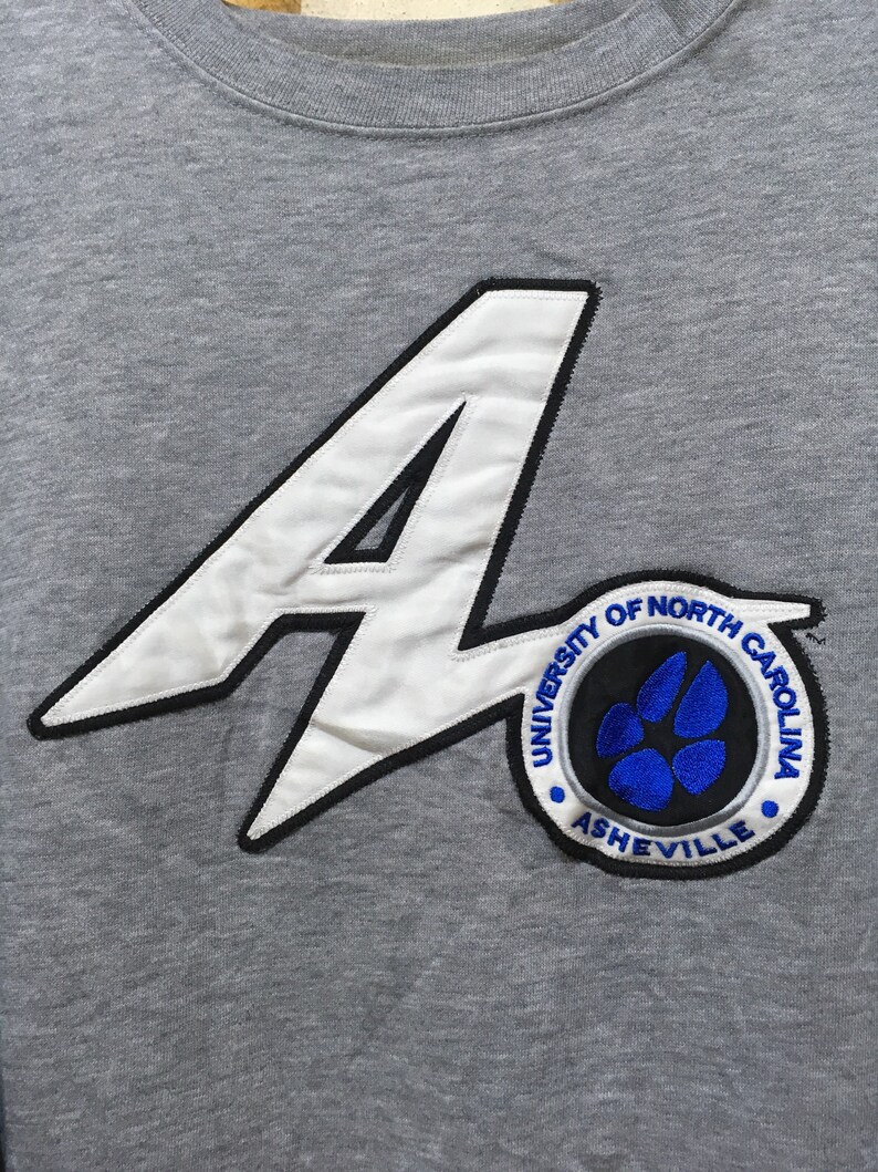 Puede incluir: Sudadera gris con una aplicaci&oacute;n de fieltro blanco de una letra A grande y un parche circular con el texto "University of North Carolina Asheville" y una huella de pata azul.