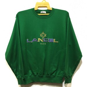 Könnte beinhalten: Grüner Sweatshirt mit einem gold- und blaustichigen gestickten Logo mit der Aufschrift "LANCEL PARIS".
