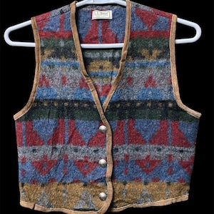 Peut inclure: Un gilet sans manches avec un motif géométrique multicolore en rouge, bleu, vert et jaune moutarde. Le vêtement présente une bordure beige le long des bords, une encolure en V et trois boutons en métal argenté. Une étiquette L.L. Bean est au col.