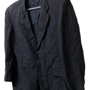 May include: A black blazer with a three-button closure, two front pockets, and a notched lapel. The jacket is hanging on a gray hanger against a white background. The fabric appears to be textured.