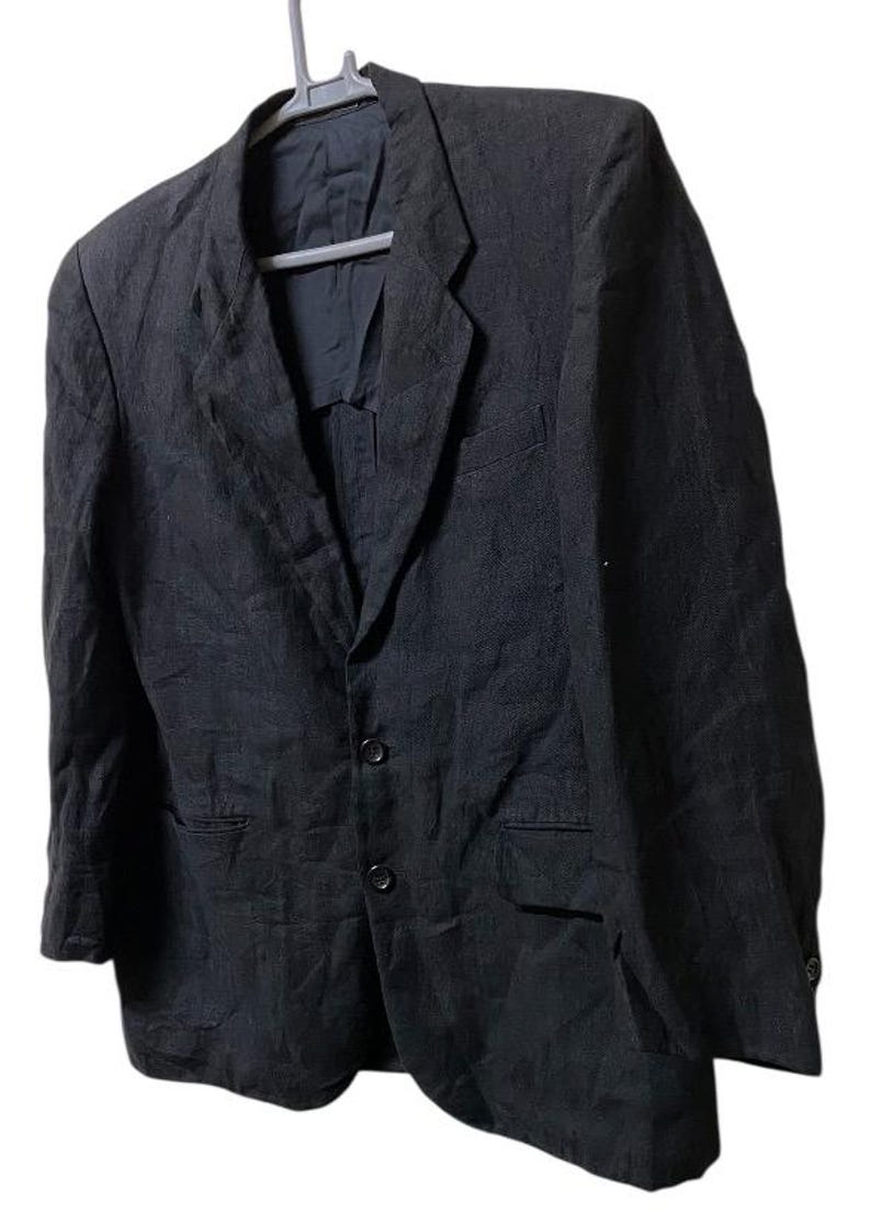 May include: A black blazer with a three-button closure, two front pockets, and a notched lapel. The jacket is hanging on a gray hanger against a white background. The fabric appears to be textured.