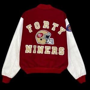 Könnte beinhalten: Rot-weiße Collegejacke mit den Worten "FORTY NINERS" in goldfarbenen Buchstaben. Die Jacke hat eine Footballhelm-Grafik und einen Aufnäher auf dem Ärmel.