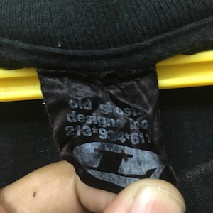 May include: Black t-shirt label with the text "old ghosts designs inc" and a phone number. The label also features a stylized logo of a top hat. The label is attached to a black t-shirt.