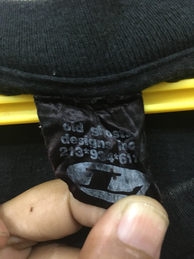 May include: Black t-shirt label with the text "old ghosts designs inc" and a phone number. The label also features a stylized logo of a top hat. The label is attached to a black t-shirt.