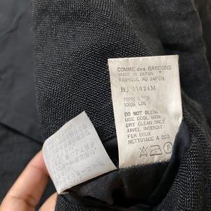 May include: Close-up of a black garment's interior, revealing a tag with text including "COMME des GARCONS MADE IN JAPAN." The tag also lists fabric composition as 100% linen and care instructions. Additional text is visible on a smaller tag.