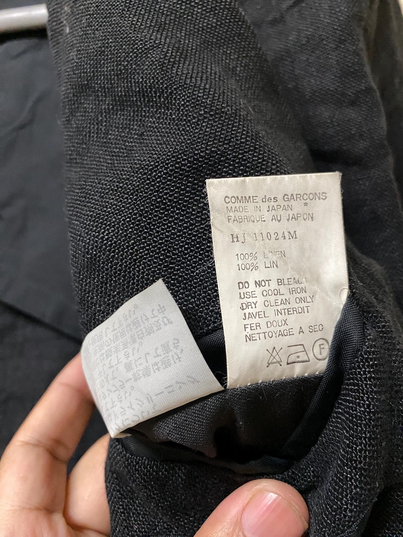 May include: Close-up of a black garment's interior, revealing a tag with text including "COMME des GARCONS MADE IN JAPAN." The tag also lists fabric composition as 100% linen and care instructions. Additional text is visible on a smaller tag.