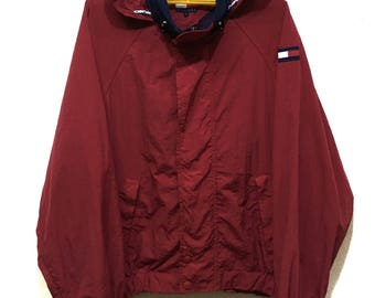 May include: A maroon hooded jacket with a navy blue and white collar, hanging on a yellow hanger. The jacket has a small logo on the sleeve. The jacket has a zip closure and two front pockets.
