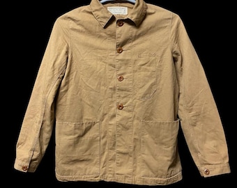 May include: A tan-colored jacket with a button-down front, a collar, and two front pockets. The jacket has a small pocket on the chest and a label near the collar. The jacket is hanging on a white hanger against a black background.