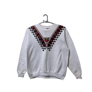 Könnte beinhalten: Ein weißer Pullover mit einem karierten und Apfel-Design auf der Vorderseite. Das Design zeigt rote Äpfel und schwarz-weiße Karos.