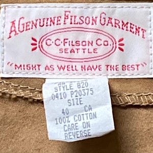 Puede incluir: Primer plano de una etiqueta de prenda beige con texto rojo "A Genuine Filson Garment" y "C.C. Filson Co. Seattle". Una etiqueta m&aacute;s peque&ntilde;a indica "Style 820, Talla 40, 100% Algod&oacute;n". La prenda es beige.