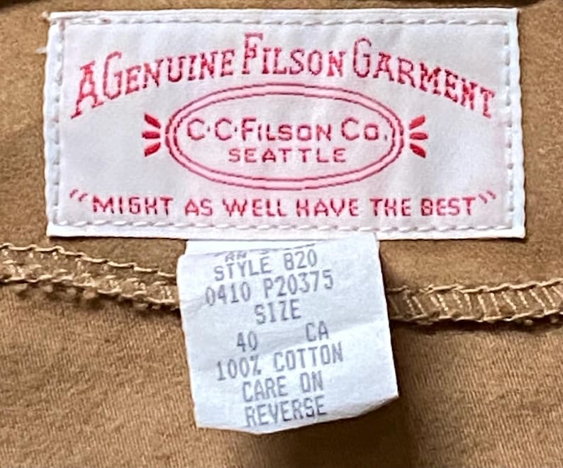 Puede incluir: Primer plano de una etiqueta de prenda beige con texto rojo "A Genuine Filson Garment" y "C.C. Filson Co. Seattle". Una etiqueta m&aacute;s peque&ntilde;a indica "Style 820, Talla 40, 100% Algod&oacute;n". La prenda es beige.