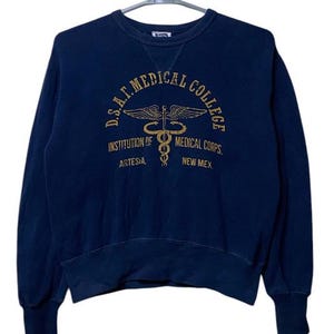 May include: A navy blue sweatshirt featuring a gold graphic of the D.S.A.F. Medical College logo. The logo depicts a caduceus, a winged staff with two snakes entwined around it, and the text "D.S.A.F. Medical College, Institution of Medical Corps, Artesia, New Mex."