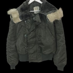 Könnte beinhalten: Dunkelgrüne Bomberjacke mit fellgefütterter Kapuze und Kragen. Die Jacke hat einen Reißverschluss, Knöpfe und gerippte Säume. Sie hängt an einem weißen Kleiderbügel.