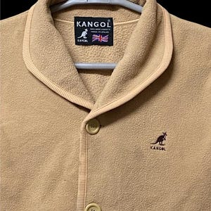 May include: A tan fleece jacket with a shawl collar and two button closure. The jacket features the Kangol logo and a small kangaroo emblem. The label includes the text "KANGOL" and the British flag.