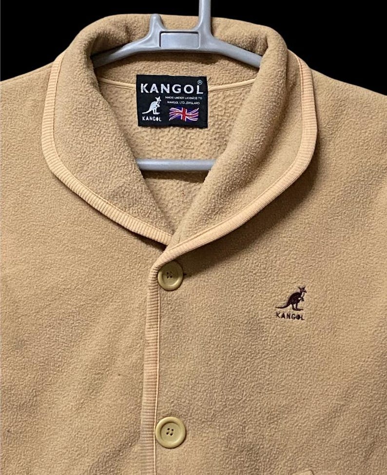 May include: A tan fleece jacket with a shawl collar and two button closure. The jacket features the Kangol logo and a small kangaroo emblem. The label includes the text "KANGOL" and the British flag.