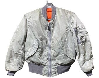 May include: A light gray bomber jacket featuring a front metal zipper closure, a ribbed gray collar, cuffs, and hem. The jacket includes two side pockets with snap button flaps, a utility pocket on the left sleeve, and a bright orange inner lining.