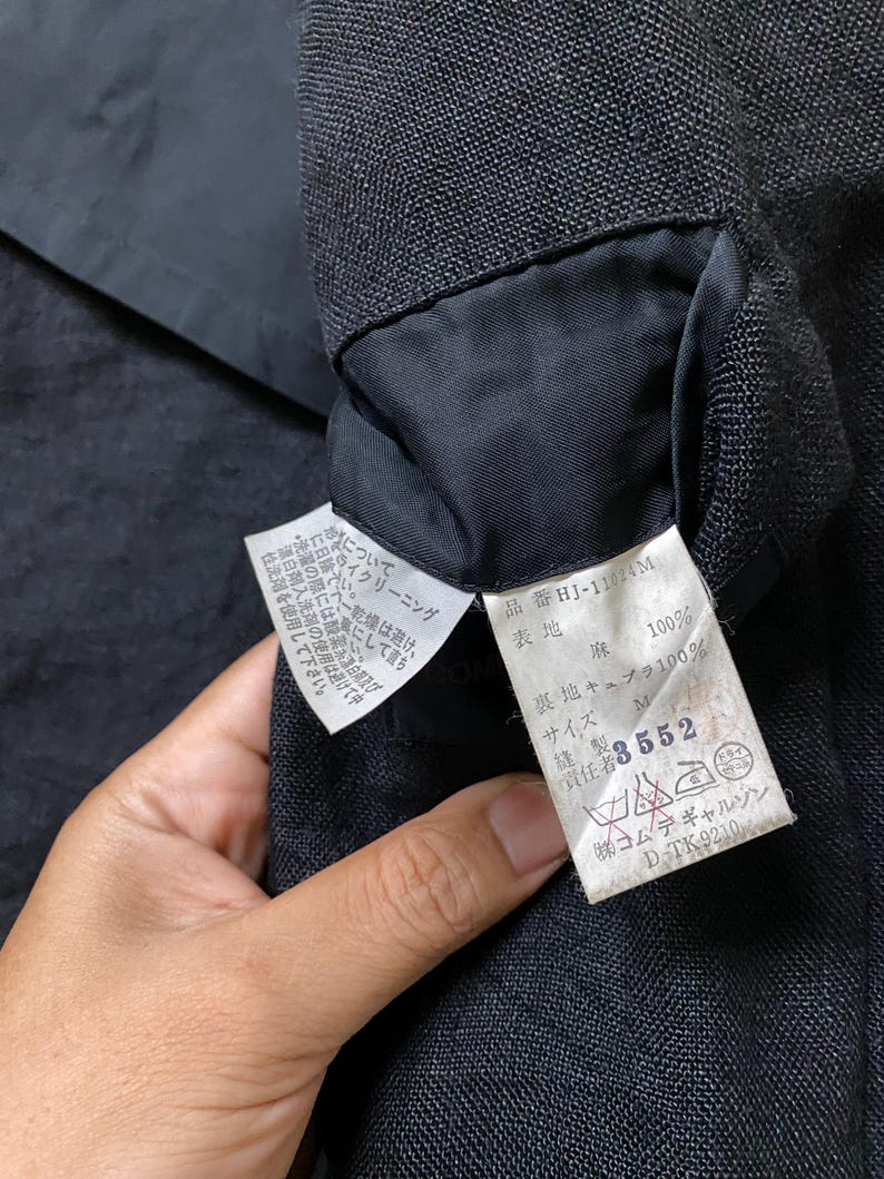 May include: Close-up of a black garment's interior, revealing a sewn-in label. The label displays Japanese text, fabric composition, and care instructions. The garment's texture is visible, with a focus on the label's details.