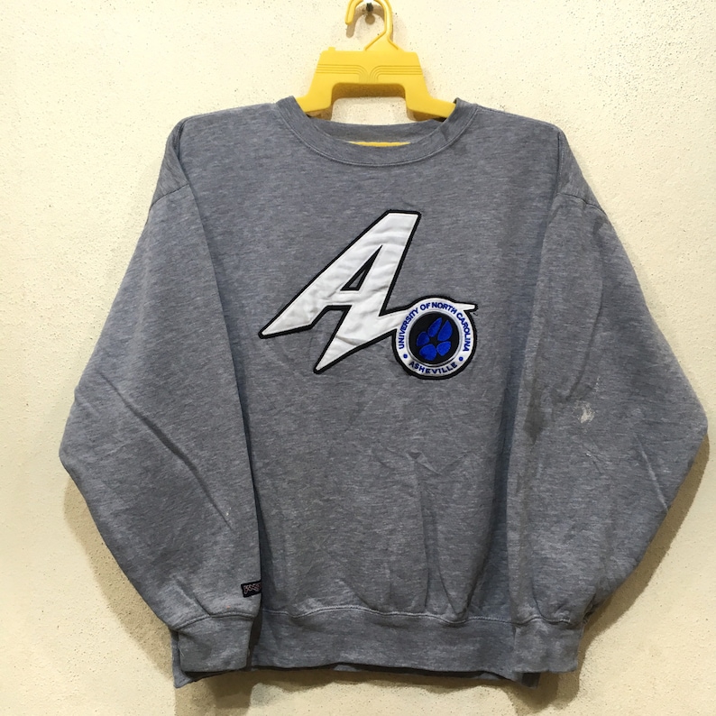 Puede incluir: Sudadera gris con un logotipo bordado en blanco y azul que dice "University of North Carolina Asheville" y una gran "A" blanca con un rayo blanco.