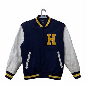 May include: A navy blue and white varsity jacket with a yellow letter "H" on the front. The sleeves are white leather and the jacket has a yellow and white striped trim.