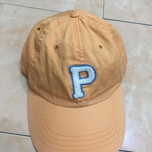 May include: A light orange baseball cap with a blue and white embroidered letter "P" on the front.