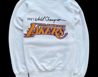 以下が含まれることがあります： オレンジと紫のロサンゼルス・レイカーズのロゴの上に「1987 World Champions」の文字が入った白いクルーネックのスウェットシャツ。スウェットシャツは長袖で、リブ編みの裾と袖口が付いています。