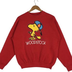 Peut inclure: Un sweat-shirt rouge avec un personnage de dessin animé jaune portant une casquette bleue et orange. Le personnage tient un bâton, avec le mot "WOODSTOCK" imprimé en blanc en dessous. Le sweat-shirt est suspendu à un cintre en bois.